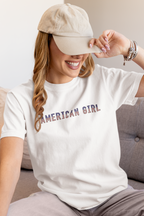 Woman wearing a white t-shirt with 'American Girl' in patriotic stars & stripes text, sitting on a couch.