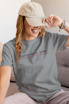 Woman wearing a granite-colored t-shirt with 'American Girl' in patriotic stars & stripes text, sitting on a couch.