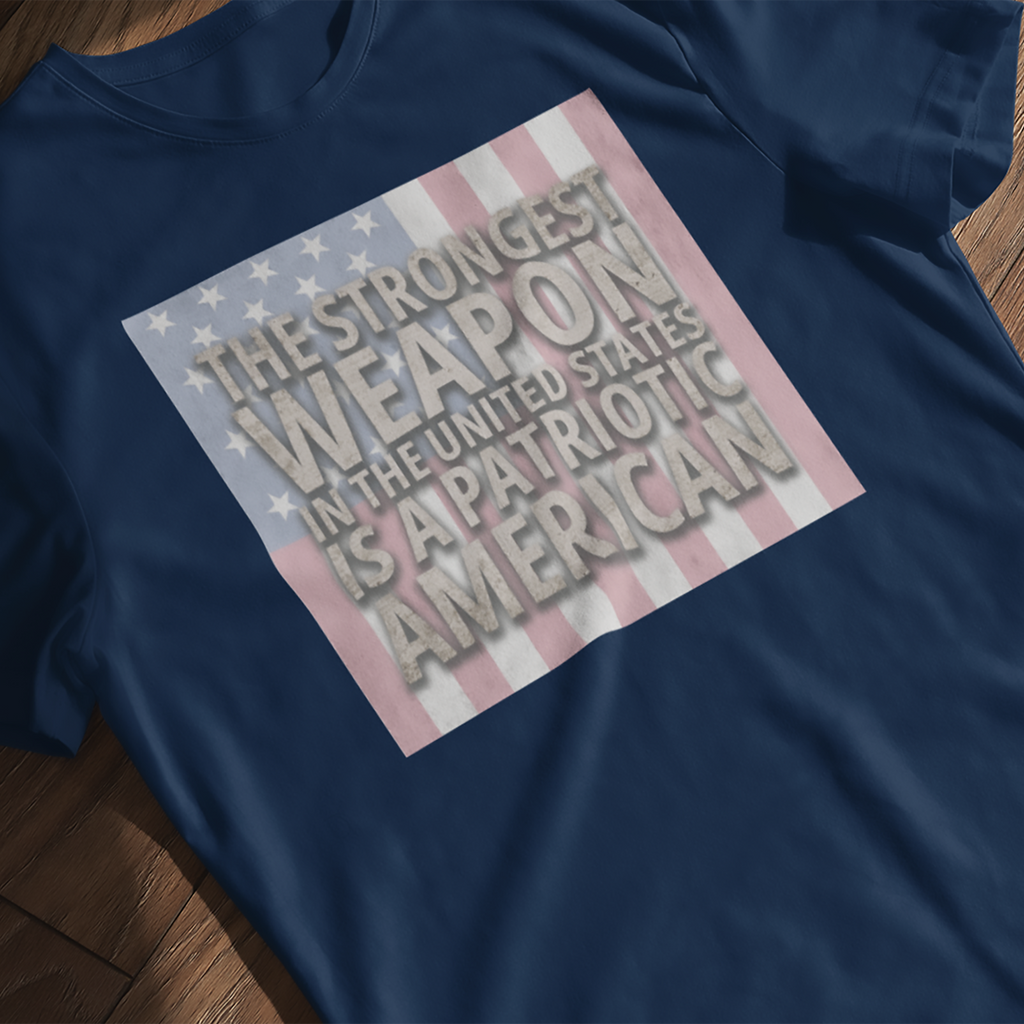 'The Strongest Weapon is a Patriotic American' t-shirt with distressed flag in navy, laying flat on a wooden surface