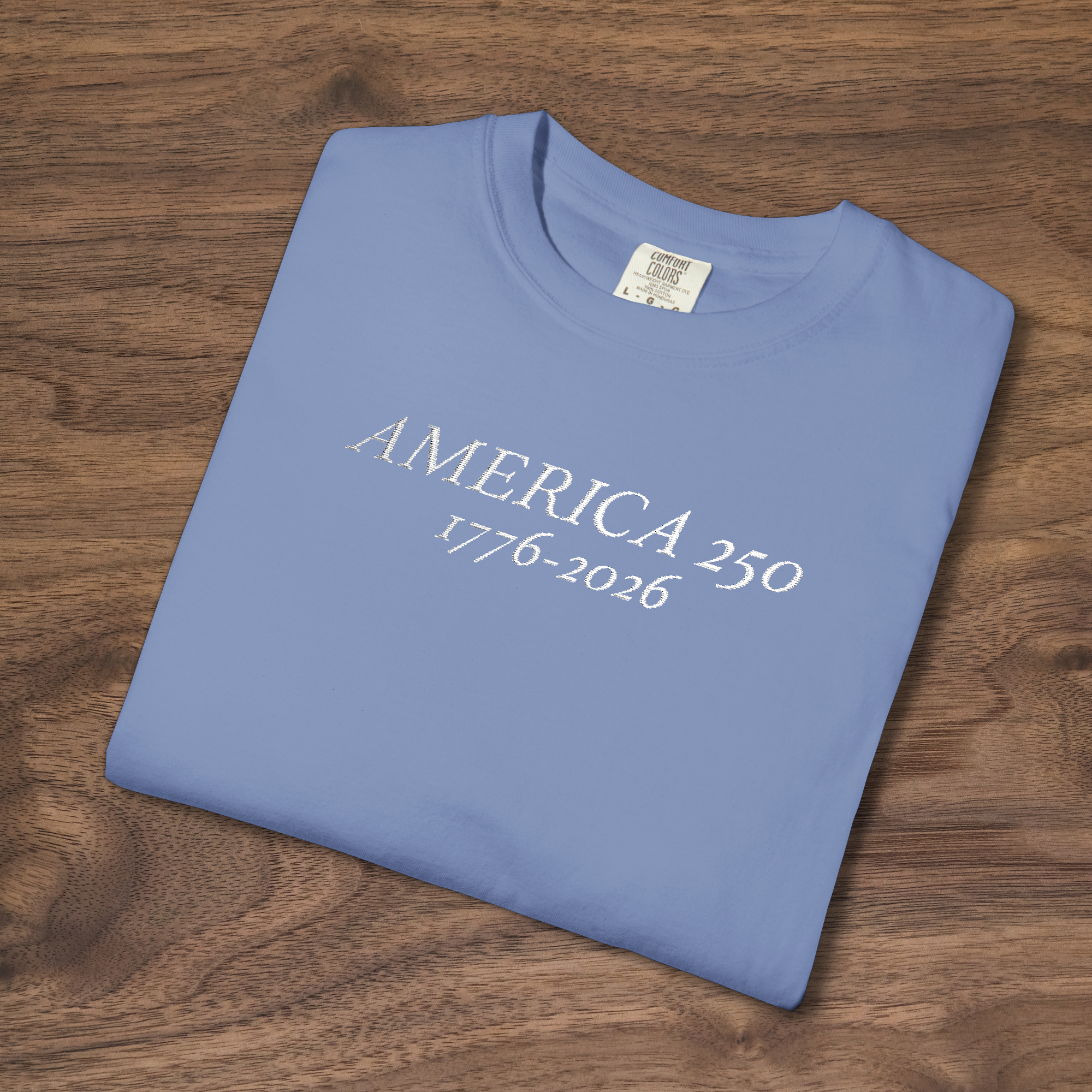 'AMERICA 250 1776-2026' embroidered on a folded light blue t-shirt laying on a wooden surface