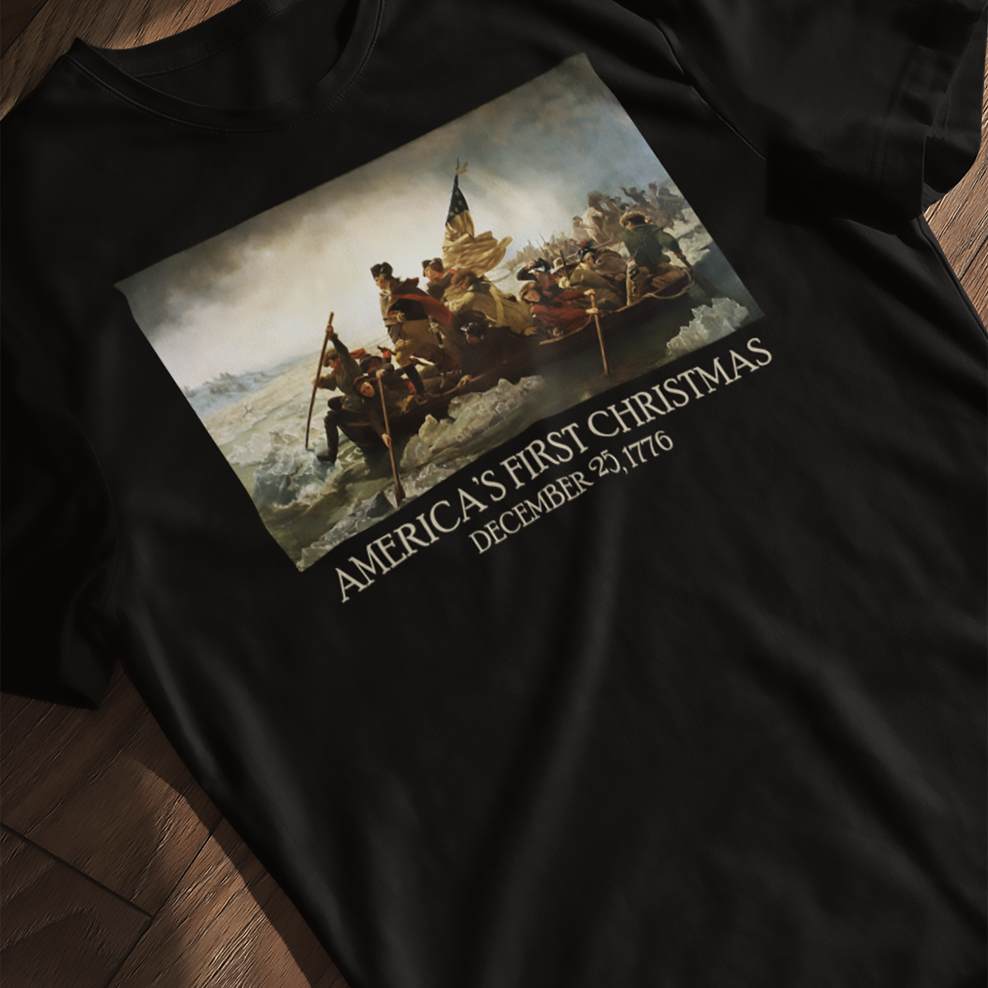 T-shirt with print of Emanuel Leutze’s 'Washington Crossing the Delaware' with 'America's First Christmas December 25, 1776' below laying flat on a wooden surface.