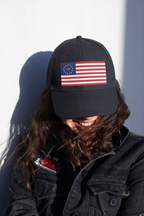 Model wearing a black cap embroidered with Betsy Ross flag