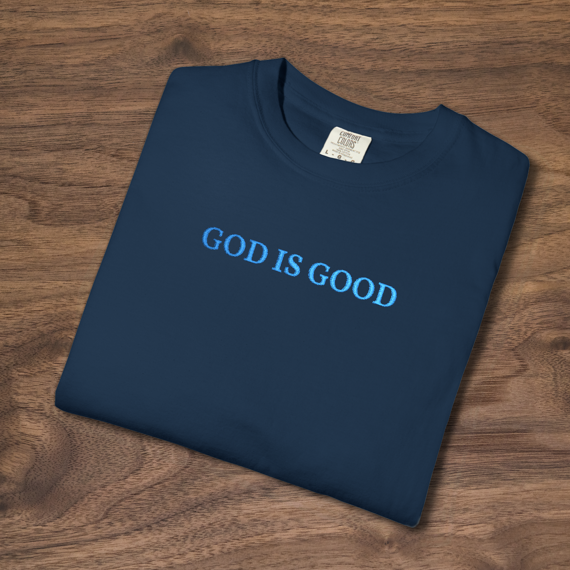 'God is Good' Christian t-shirt embroidered with cornflower blue thread, folded, laying on a wooden surface