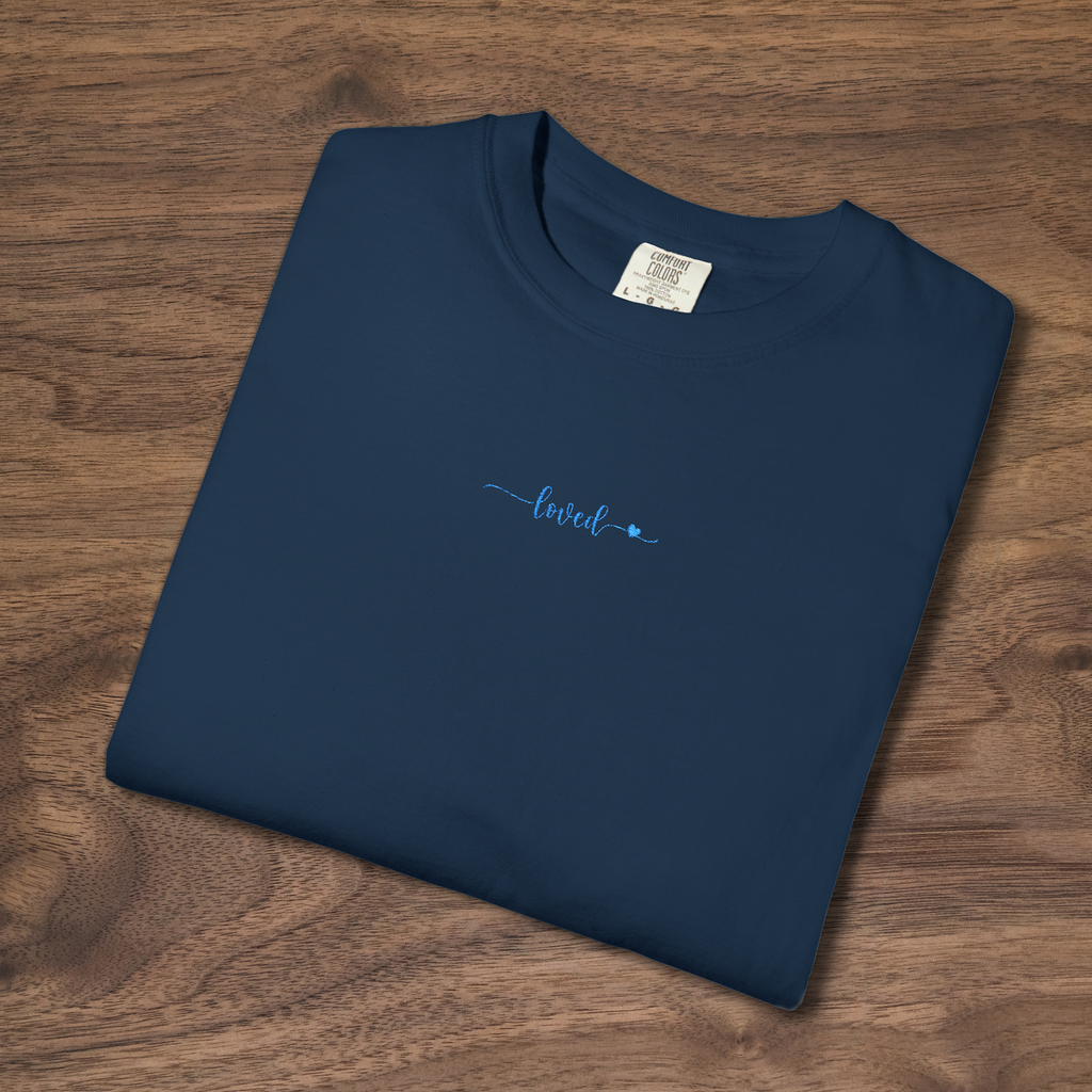 t-shirt with 'loved' plus a tiny heart embroidered with cornflower blue thread, folded, laying on a wooden surface